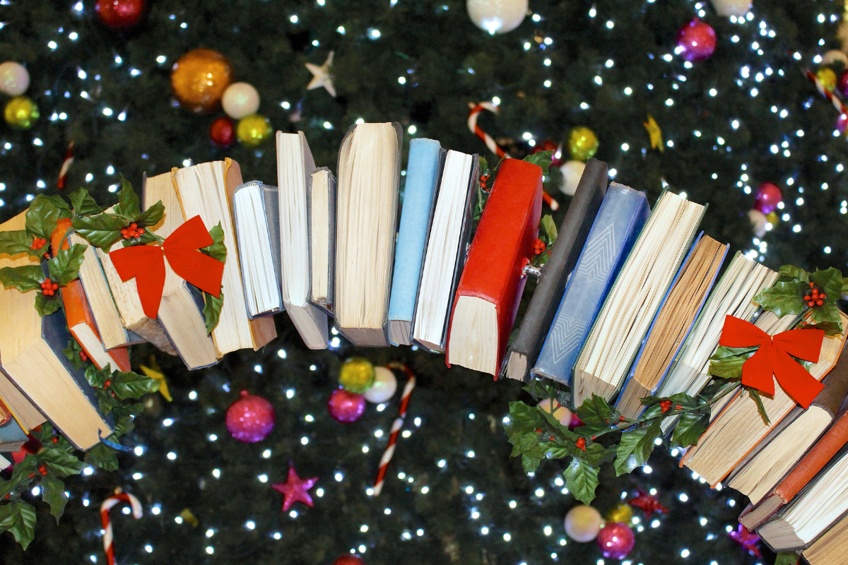 Dekoracyjna girlanda z otwartymi książkami ozdobiona gałązkami ostrokrzewu i czerwonymi kokardami, zawieszona na tle świątecznej choinki z kolorowymi bombkami i migoczącymi światełkami.